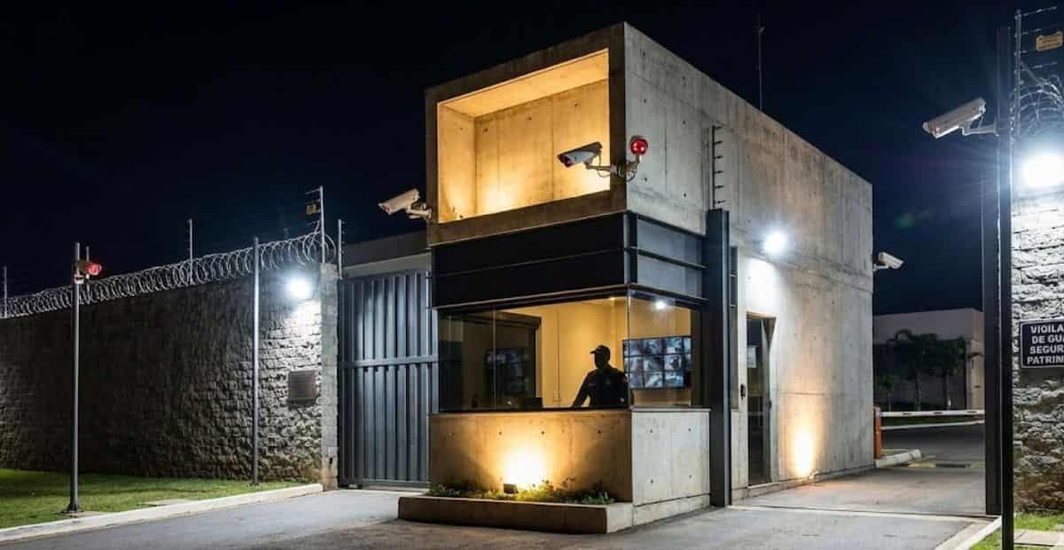 Foto noturna de uma guarita de segurança patrimonial moderna e blindada, com muros altos, cerca elétrica e múltiplas câmeras de vigilância protegendo um perímetro.